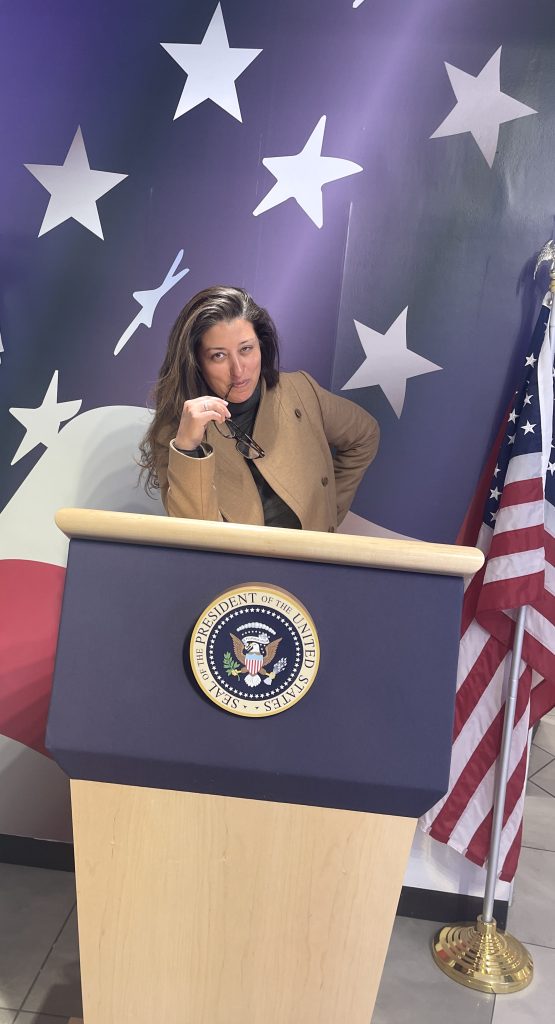 Woman standing in front of a podium that has the flag of the USA draped behind and has the Presidential Seal on the front of the podium