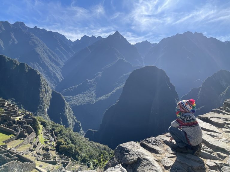 Child seated on cliff edge staring across a deep valley at the ruins of Machu Pichu against a tall mountain backdrop