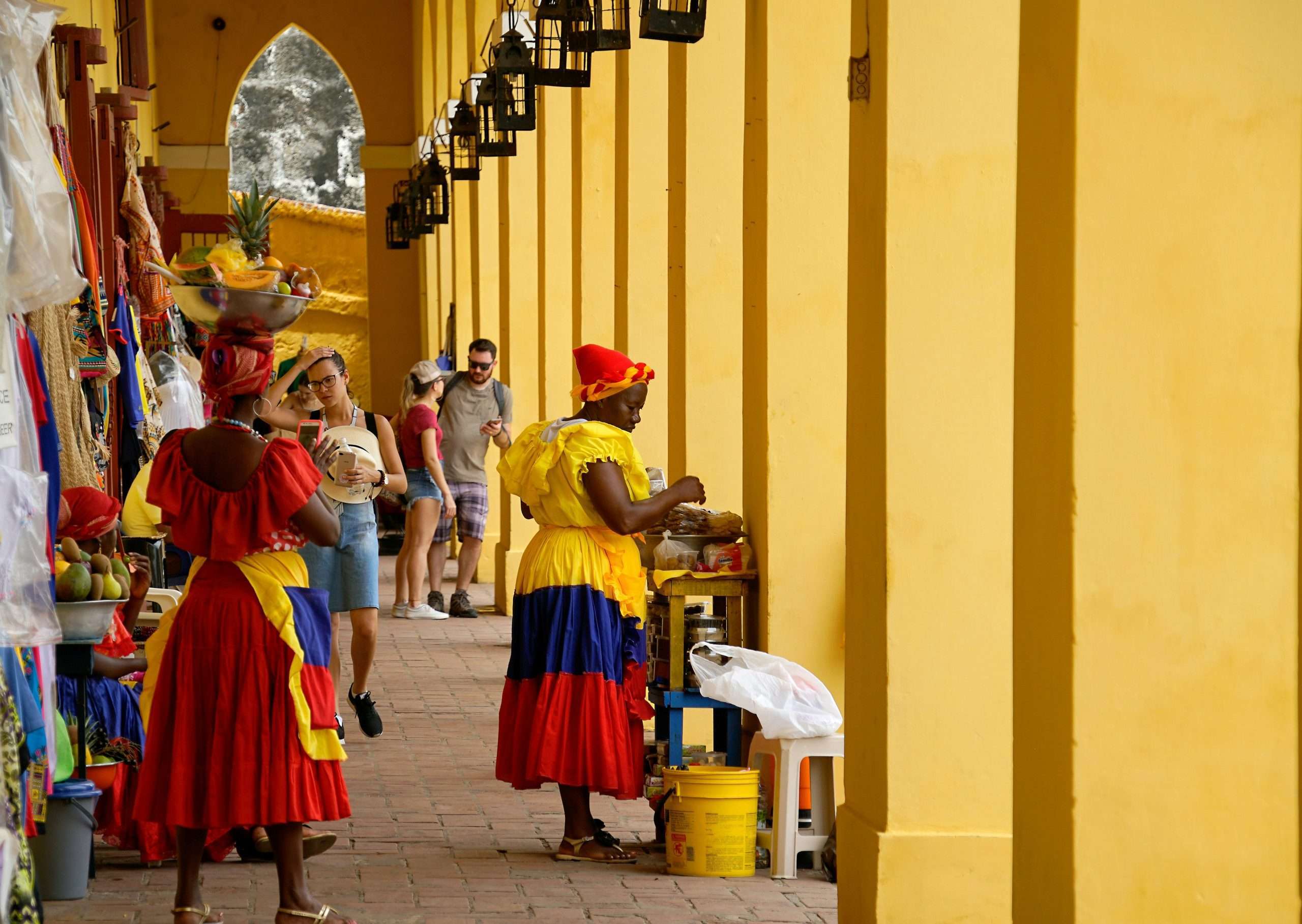 Women along brick pathway under a bright yellow building entrance dressed in traditional Colombian colors selling handicrafts | Photo by Ricardo Gomez Angel on Unsplash