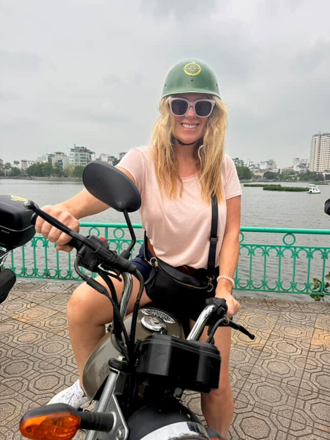 Caucasian blonde woman sitting on a motorcycle and holding the handlebars, wearing a helmet and sunglasses, smiling broadly, in front of a waterway