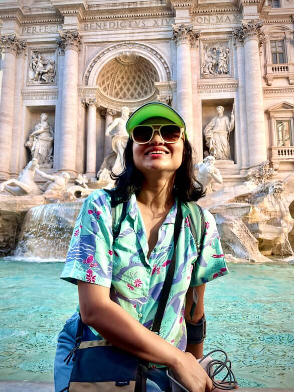 South Asian woman sitting alone smiling with sunglasses and a bright hat in front of the marble sculpture Trevi Fountain in Rome