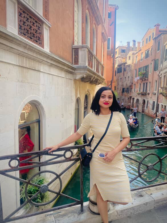 South Asian woman standing alone on a footbridge over a small waterway with hand-rowed boats in the background in Venice, Italy