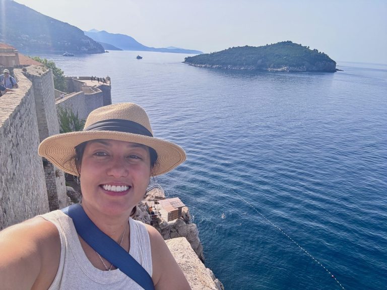 Woman squinting into the sun with a broad-brimmed hat as she leans out over an ancient stone wall into expansive ocean behind