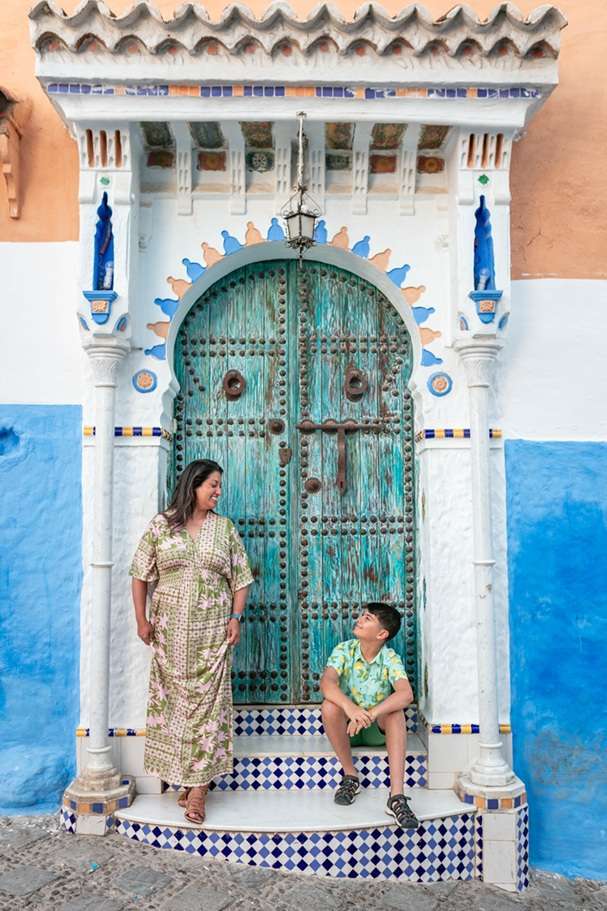 Woman standing in a flowing dress and young boy seated, both in a blue and white elaborately painted doorframe in the mosaic tiled streets of Chefchaouen, Morocco.