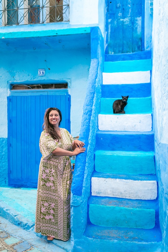 Woman standing and smiling by a concrete set of brightly blue painted stairs with a black cat seated on the stairs