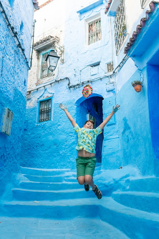 Young laughing boy jumping in the air in front of bright blue painted concrete stairs in Chefchaouen