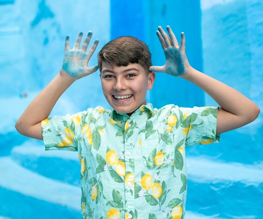 Smiling young boy wearing a pale green shirt with bright yellow lemons making a silly face as he stands in front of bright blue painted concrete stairs showing the blue paint on his hands
