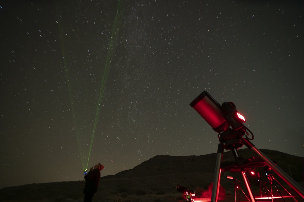 Telescope glowing red under the Milky Way with green laser beams pointing toward the stars in Joshua Tree.
