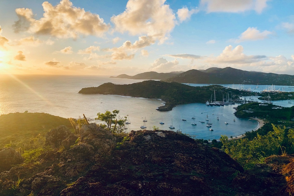 Sunset view over English Harbour in Antigua, with sailboats anchored in the turquoise bay and golden light illuminating the surrounding green hills and coastline.