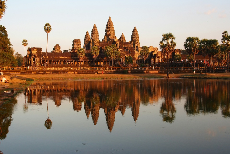 The ancient temple complex of Angkor Wat in Cambodia reflected in a calm lotus pond at sunrise, surrounded by palm trees and golden morning light.