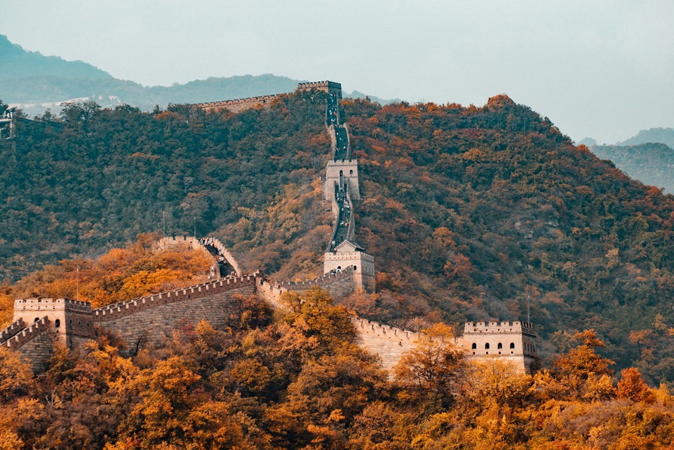 The Great Wall of China winding over forested mountains covered in autumn foliage, with watchtowers stretching into the misty distance.