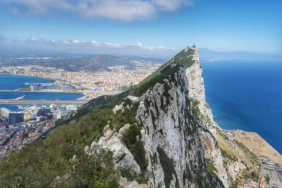View of the Rock of Gibraltar rising above the city and the Mediterranean Sea, with Spain visible in the distance under a clear blue sky.