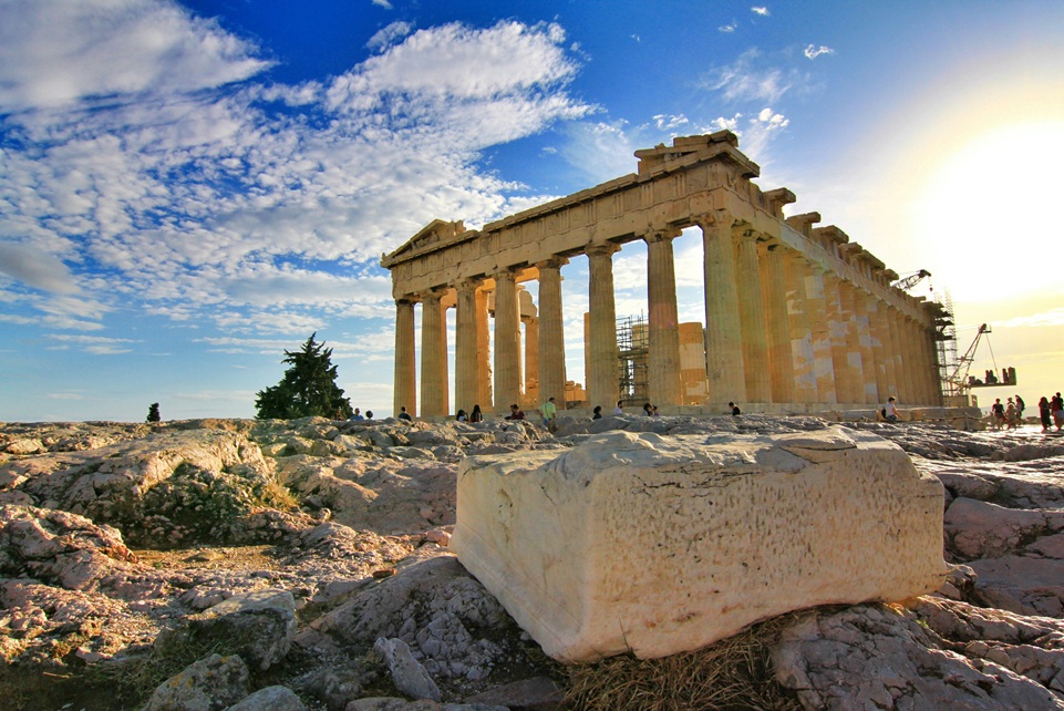 The Parthenon atop the Acropolis in Athens, Greece, with visitors exploring the ancient marble ruins under a bright blue sky and scattered clouds.
