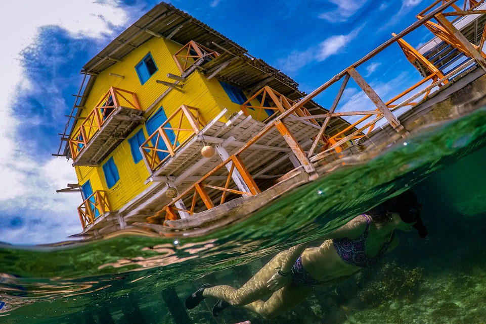Snorkeler swimming below a bright yellow overwater guesthouse in Grenada, with clear turquoise water and coral reef visible beneath the surface.