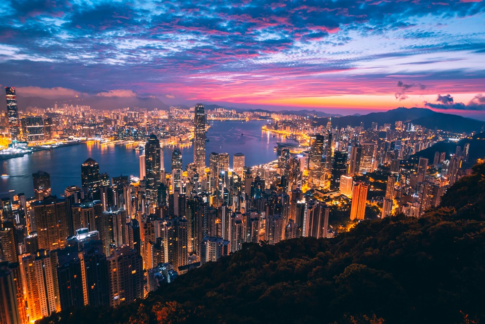 Panoramic view of Hong Kong’s skyline at sunset, with illuminated skyscrapers reflecting on Victoria Harbour and colorful clouds glowing above the city.