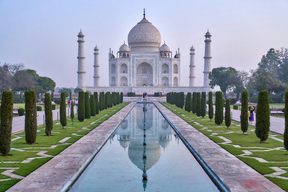 The Taj Mahal in Agra, India, beautifully reflected in its long marble pool, surrounded by manicured gardens and visitors strolling along the path at sunrise.