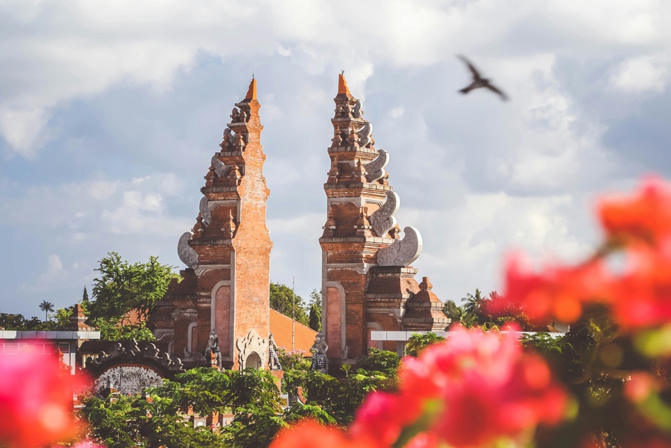 Traditional split gateway temple in Bali, Indonesia, framed by bright tropical flowers and a flying bird against a cloudy sky.