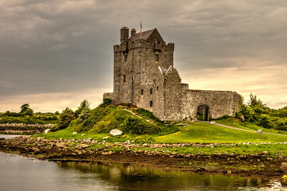 Medieval stone castle on a grassy hill beside a calm river in Ireland, with the Irish flag flying against a cloudy golden sky.