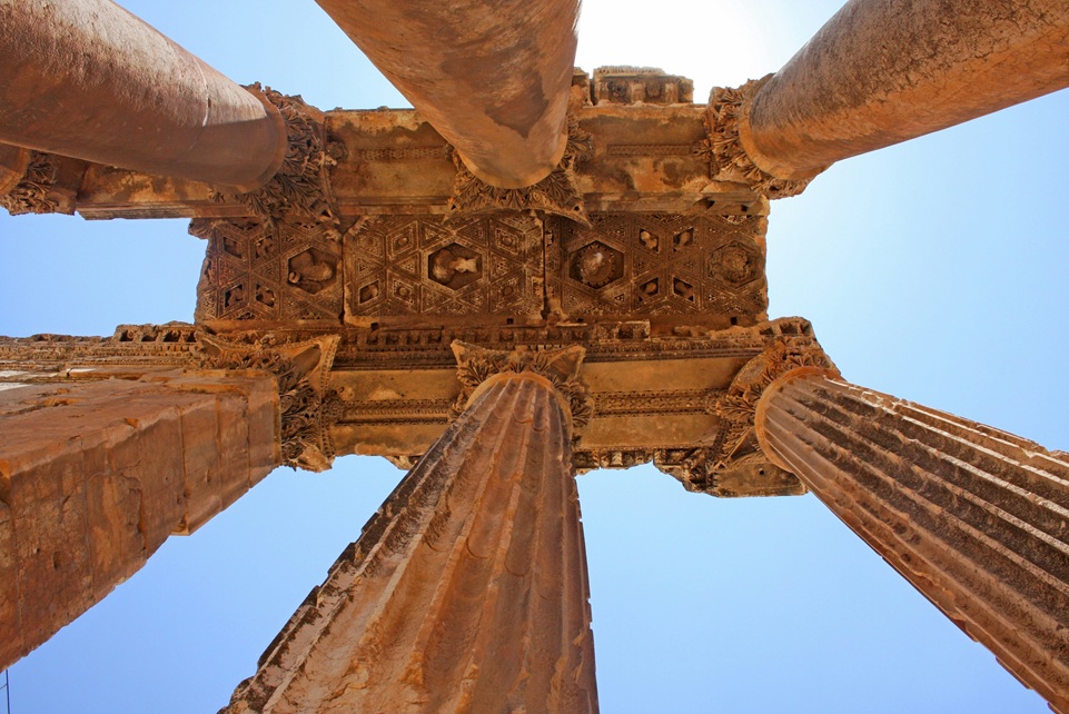 Upward view of the ancient Roman columns of Baalbek Temple in Lebanon, showing intricate carvings and weathered stone against a bright blue sky.