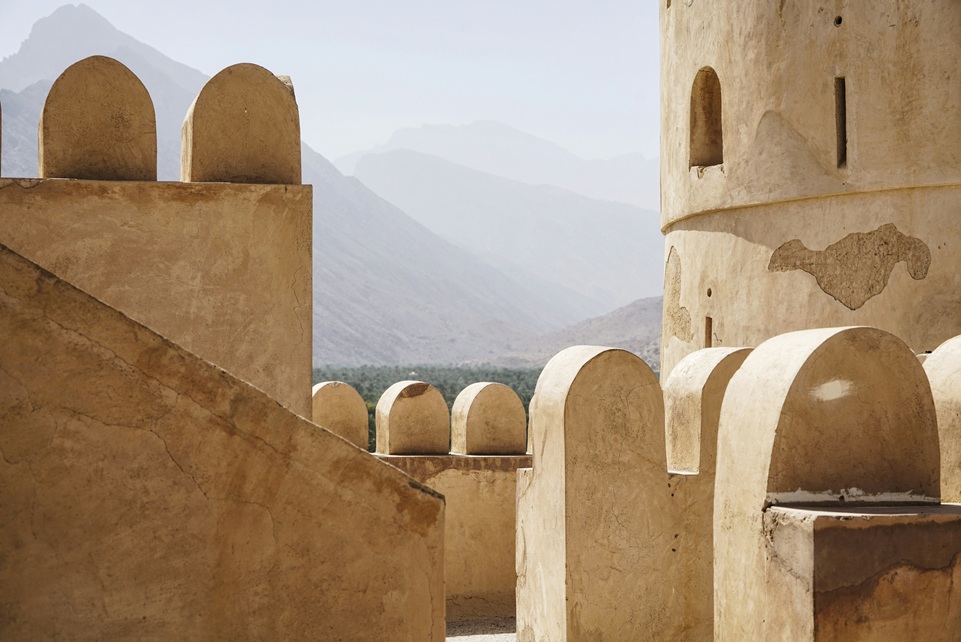 Traditional desert fortress in Oman with sand-colored stone walls and rounded battlements, set against a backdrop of hazy mountains and palm-filled valleys.