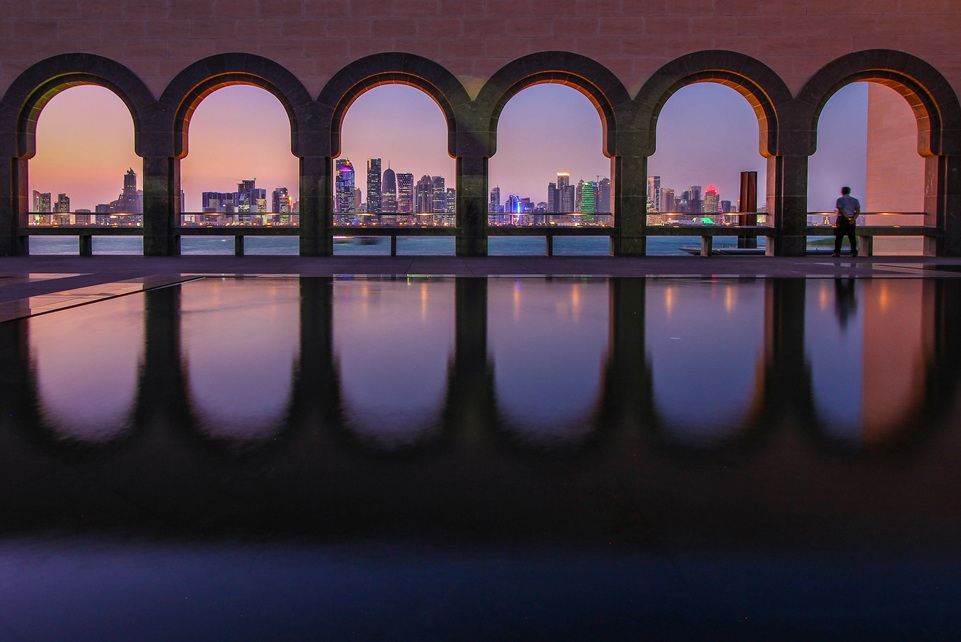 Doha skyline at sunset viewed through the arched windows of the Museum of Islamic Art in Qatar, with reflections shimmering in the courtyard pool.