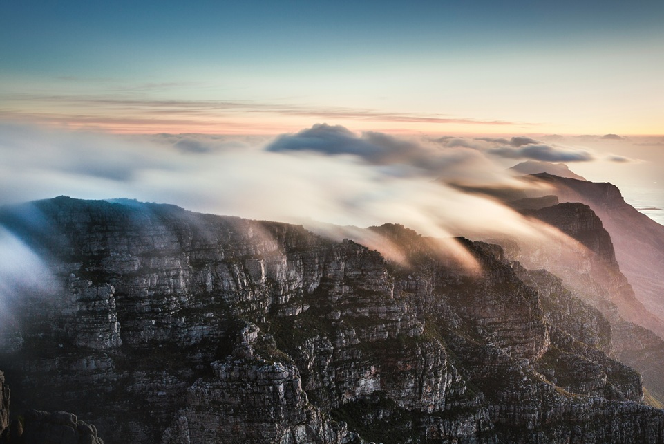 Sunrise clouds rolling over Table Mountain in Cape Town, South Africa, with golden light illuminating the rugged cliffs and coastline below.
