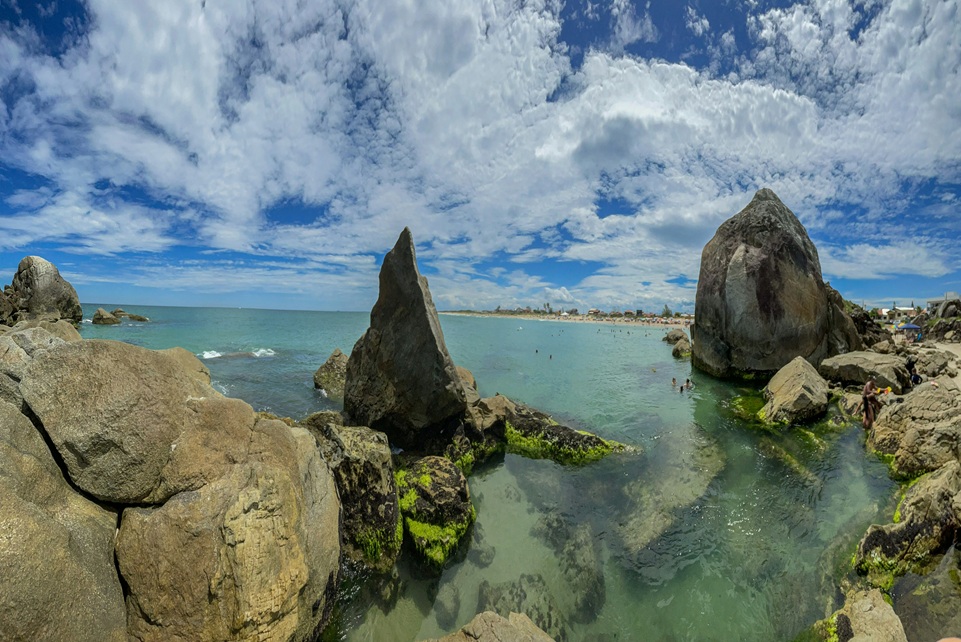 Rocky coastline and clear turquoise water in St. Vincent and the Grenadines, with people swimming near large boulders under a bright blue tropical sky.