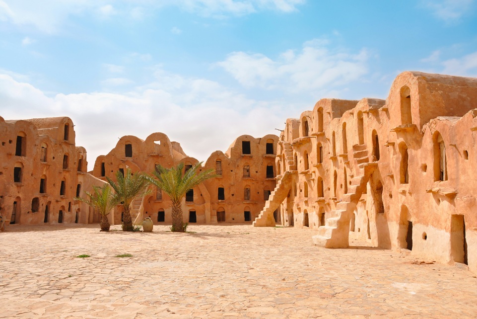 Traditional Berber granary architecture in the Ksour region of southern Tunisia, with earthen buildings, arched doorways, and palm trees under a bright desert sky.
