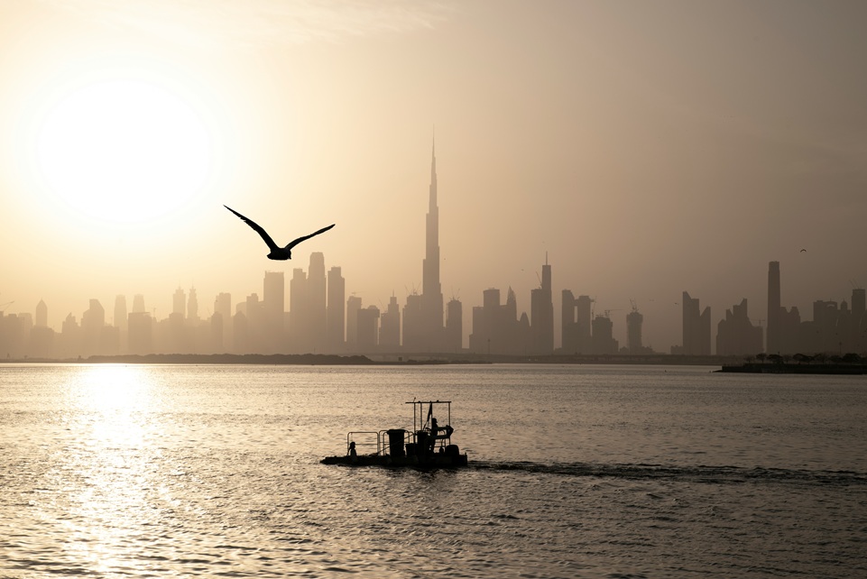 Silhouette of Dubai’s skyline at sunset with the Burj Khalifa towering above the city, a bird soaring overhead, and a small boat crossing the calm waters of the UAE coast.