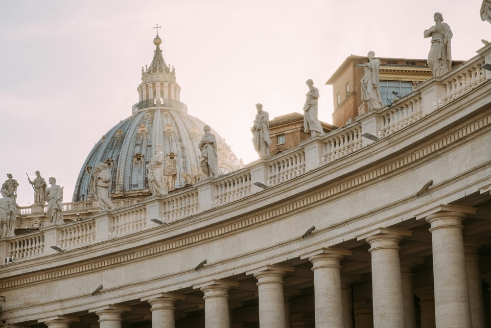 St. Peter’s Basilica dome and statues glowing in the morning light above the colonnade of St. Peter’s Square in Vatican City.