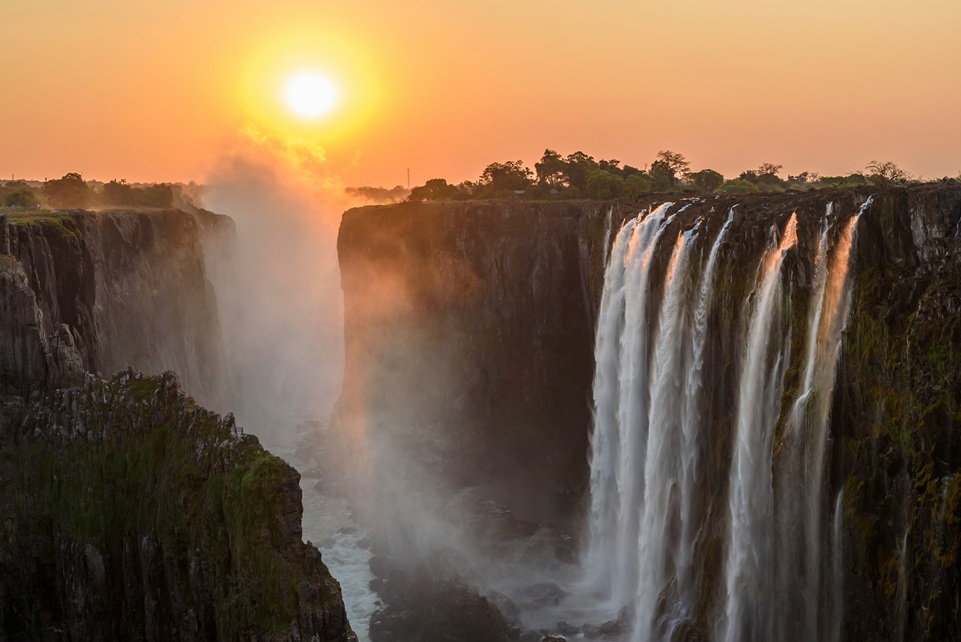 Victoria Falls at sunset on the Zimbabwe side, with golden light illuminating the mist and cascading water between steep cliffs.