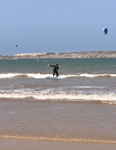Teen surfer riding a wave during a beginner surf lesson in Essaouira, Morocco, with kitesurfers visible along the Atlantic coast.